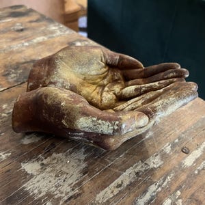 May include: A decorative sculpture shaped like two cupped hands joined together. The object features a weathered, distressed finish with mottled shades of brown, tan, and muted gold, creating a textured appearance across the palms and fingers.