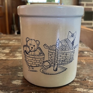 May include: A cylindrical beige stoneware crock featuring a dark blue line drawing of a teddy bear in a basket, a cat in a basket, and a tall candle. The illustration includes the text '©94 K.HUGHES' printed near the base of the basket.
