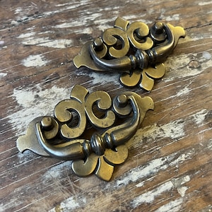May include: Two antique-style brass drawer pulls featuring a decorative backplate with a scrolled, symmetrical design and a curved, swinging handle. The metal has a darkened, aged patina with visible highlights on the raised surfaces.
