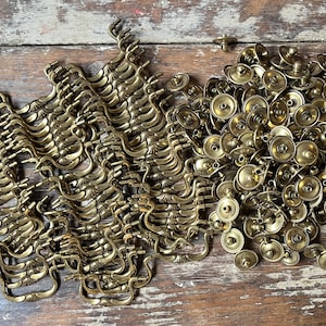 May include: A large collection of brass-toned metal furniture hardware, including numerous curved swing-style drawer pulls and circular backplates with central mounting holes, arranged in a dense pile of individual components.