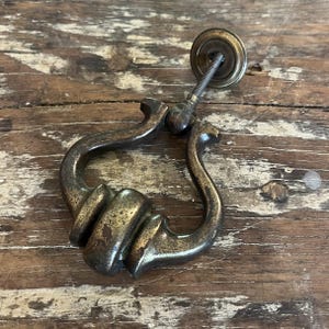 May include: A vintage-style brass drawer pull featuring a curved, drop-handle design with a weathered, dark patina. The hardware includes a circular mounting plate and a threaded metal bolt extending from the back for secure attachment to furniture.