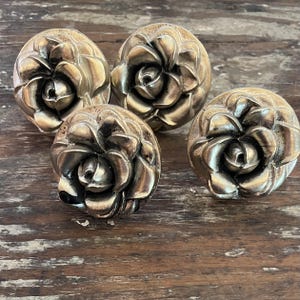 May include: A set of four circular cabinet knobs, each featuring a three-dimensional rose flower design. The knobs are finished in a metallic antique brass tone with dark highlights in the crevices of the petals to emphasize the floral shape.