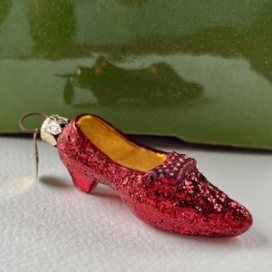 May include: A red glittery shoe ornament with a gold interior and a small metal loop at the heel for hanging. The shoe features a low heel and a decorative bow detail on the front, covered in sparkling red material throughout the exterior.