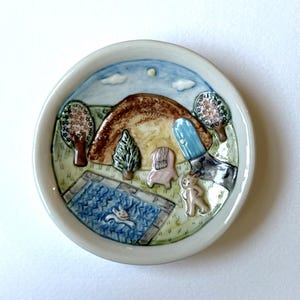 May include: A round ceramic dish featuring a raised, hand-painted landscape scene. The design includes a brown hill, a blue swimming pool with a figure swimming, a small chair, a blue door, two trees, and a figure standing on green grass.