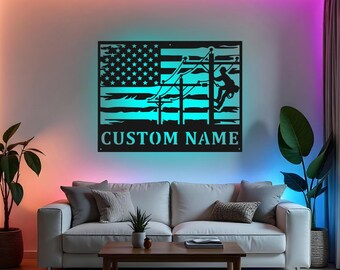 May include: Black metal wall art featuring an American flag design with a lineman climbing a utility pole. The artwork includes the text "CUSTOM NAME" at the bottom. The piece is illuminated with a blue glow, creating a striking visual.