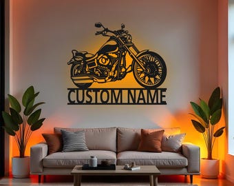 May include: Black metal motorcycle wall art with the text "CUSTOM NAME" below the motorcycle. The motorcycle is illuminated with a warm orange glow, mounted on a light-colored wall above a beige sofa.