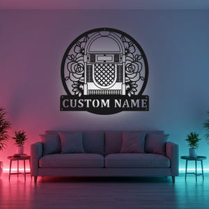 Op de afbeelding: Zwarte metalen wandkunst met een jukebox-ontwerp met bloemmotieven. De jukebox is geplaatst in een cirkelvormig frame, en de woorden "CUSTOM NAME" worden weergegeven op een rechthoekige banner eronder. De kunst is aan een muur boven een grijze bank bevestigd.