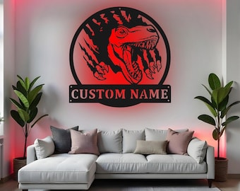 May include: A black metal wall art piece featuring a red dinosaur design with the text "CUSTOM NAME". The dinosaur is inside a circle with claw marks. The art is mounted above a white couch.