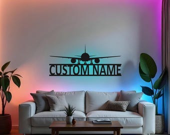 Puede incluir: Una silueta de avión de metal negro con el texto "CUSTOM NAME" debajo. El avión está montado en una pared con un fondo blanco y luces LED de colores. Un sofá con cojines y una mesa de café están en primer plano.