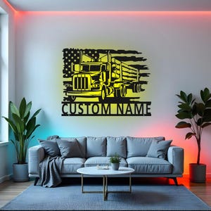 Op de afbeelding: Een zwart en geel metalen wandkunstwerk met een vrachtwagen die boomstammen vervoert, tegen een gestileerde Amerikaanse vlag. De tekst "CUSTOM NAME" staat onder de vrachtwagen. Het kunstwerk is op een witte muur gemonteerd.