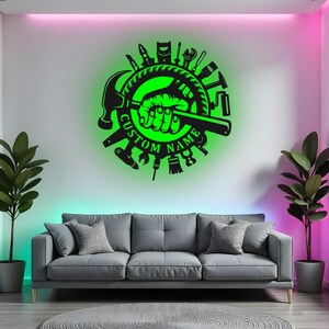 Può includere: Un'opera d'arte da parete in metallo nero con un design illuminato di verde che raffigura una mano che tiene una chiave inglese, circondata da strumenti. Il design circolare include le parole "CUSTOM NAME". L'opera è montata su un muro bianco sopra un divano grigio.