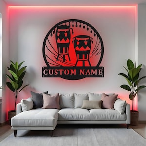 Puede incluir: Arte mural de metal negro con dos tambores dentro de un marco circular con luces de cadena. Debajo de los tambores, un letrero rectangular que dice "CUSTOM NAME". La obra de arte está montada en una pared blanca sobre un sofá seccional blanco.