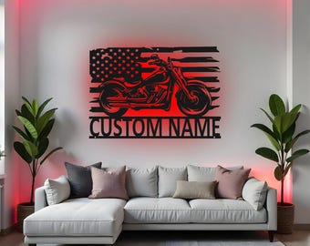 May include: A black metal motorcycle silhouette against a red-lit American flag design. The artwork includes the text "CUSTOM NAME" below the motorcycle. The wall art is displayed above a white sectional sofa with decorative pillows.