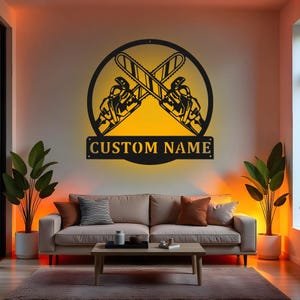 May include: A black metal wall art piece featuring crossed chainsaws within a circular frame. The text "CUSTOM NAME" is displayed on a rectangular plaque below. The art is illuminated with a warm yellow glow, creating a striking contrast against the wall.