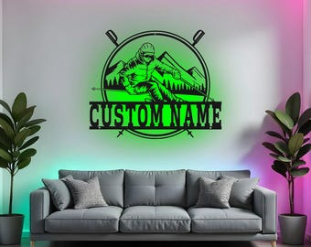 Op de afbeelding: Een zwart metalen wandkunstwerk met een skiër in een groene omtrek, tegen een bergachtergrond. Het ontwerp bevat de tekst "CUSTOM NAME" onder de skiër, perfect voor personalisatie. Het kunstwerk is op een witte muur gemonteerd.