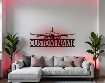 Puede incluir: Arte de pared de avión de metal negro con el texto "CUSTOM NAME" debajo. El arte se muestra en una pared blanca con una tira de luz roja encima. Un sofá seccional blanco está en primer plano.