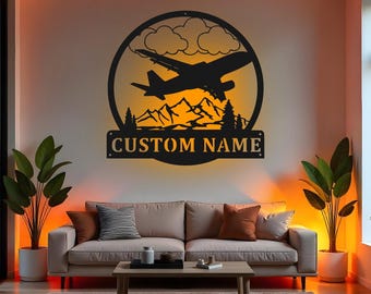 Puede incluir: Arte de pared de metal negro con la silueta de un avión dentro de un marco circular, con montañas, nubes y árboles. El texto "CUSTOM NAME" se muestra debajo. El arte está montado en una pared de color claro, encima de un sofá.