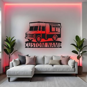 Könnte beinhalten: Schwarze Metall-Wandkunst eines Imbisswagens mit dem Text "CUSTOM NAME" darunter. Der Imbisswagen hat einen roten Schein dahinter und ist an einer weißen Wand über einem weißen Ecksofa angebracht. Zwei Topfpflanzen flankieren das Sofa.
