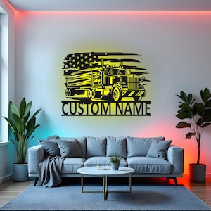 Op de afbeelding: Zwart metalen wandkunst met een silhouet van een vrachtwagen tegen een geel en zwart Amerikaans vlagontwerp. De tekst "CUSTOM NAME" staat eronder. Het kunstwerk hangt aan een witte muur boven een grijze bank.