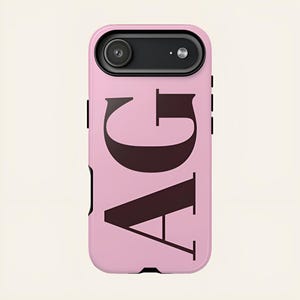 May include: A light pink smartphone case featuring the large, dark brown capital letters AG printed vertically on the back. The case has a black camera housing at the top with a circular lens and a small flash opening, and black side buttons.