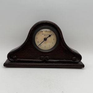May include: A dark brown wooden mantel clock with a circular clock face. The clock face has Roman numerals and the text "Regency Clock Makers 1703". The clock has a curved top and decorative scrollwork.