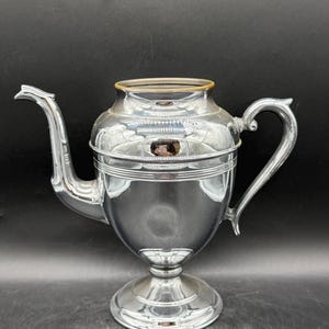 May include: A silver-colored, vintage teapot with a curved spout and handle. The teapot has a bulbous body and sits on a round base. The top rim is accented with a gold-colored band. The teapot is made of metal and has a reflective surface.