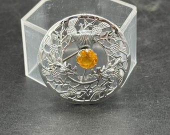 May include: Silver-toned brooch with a circular design featuring thistle motifs. The brooch has an orange-yellow gemstone at its center. The brooch is set against a clear, geometric background.