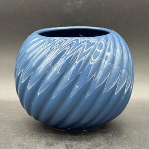 Op de afbeelding: Een blauwe keramische vaas met een ronde vorm en een getextureerd, wervelend patroon. De vaas heeft een brede opening en een glanzende afwerking. De kleur is een gedempt blauw, en het ontwerp doet denken aan een spiraal. De vaas is geschikt voor woondecoratie.
