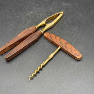 May include: A nutcracker and corkscrew set. The nutcracker has a brass head and wooden handles. The corkscrew has a brass screw and a wooden handle. The set is on a dark surface.