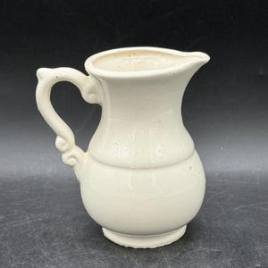 May include: Cream-coloured ceramic jug with a curved handle and a pouring spout. The jug has a rounded body with a horizontal band around the middle. The handle features decorative swirls. The jug is likely for serving liquids.