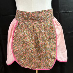 Puede incluir: Un delantal de media cintura de estilo vintage con un estampado floral sobre un fondo marrón. El delantal tiene paneles laterales rosas y un ribete rosa. El delantal se muestra en un maniquí.