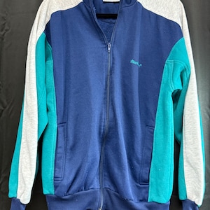 Könnte beinhalten: Eine marineblaue Vintage-Trainingsjacke mit Reißverschluss und türkisfarbenen und grauen Farbakzenten. Die Jacke hat einen hohen Kragen, Vordertaschen und das gestickte Wort "Mach" auf der Brust. Die Jacke hängt an einem schwarzen Kleiderbügel.