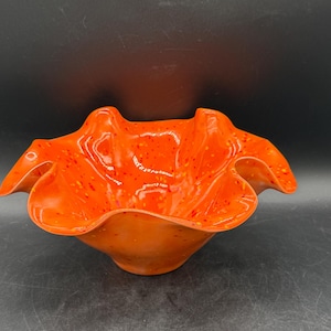 Può includere: Una ciotola in ceramica arancione brillante con un bordo increspato. La ciotola ha una finitura lucida ed è macchiata di puntini rossi e gialli. La forma della ciotola è organica e fluida, suggerendo una qualità artigianale. La ciotola è su uno sfondo scuro.