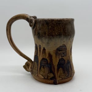 May include: Handmade ceramic mug with a unique, textured design. The mug features a brown and tan color scheme with dark brown drips and a curved handle. The mug is suitable for coffee, tea, or other beverages.