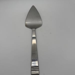 Può includere: Una paletta per torte in acciaio inossidabile con una lama triangolare e un lungo manico rettangolare. Il manico presenta tre linee orizzontali vicino all'estremità. Progettato per tagliare e servire torte.