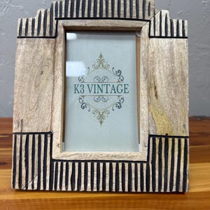 May include: Wooden picture frame with a rectangular opening and a decorative top. The frame is made of light-colored wood with black vertical stripes. The text "K3 VINTAGE" is visible on the glass.