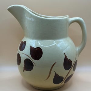 Może przedstawiać: Kremowy dzbanek ceramiczny o zaokrąglonym korpusie, zakrzywionym uchwycie i szerokim dzióbku. Zewnętrzna powierzchnia ozdobiona jest ręcznie malowanymi ciemnobrązowymi motywami liści na cienkich, wijących się łodygach wokół dolnej części.