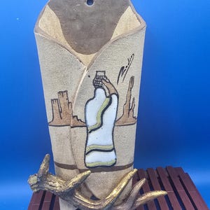 May include: A beige ceramic wall pocket with a textured surface featuring a painted illustration of a figure carrying a basket on their head in a desert landscape. A gold-toned antler-shaped accent is attached to the front lower section.
