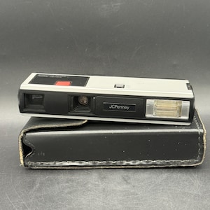 May include: A vintage JCPenney Pocket 110 camera, silver and black, resting on a black leather case. The camera has a red button and a flash. The JCPenney logo is visible on the front.