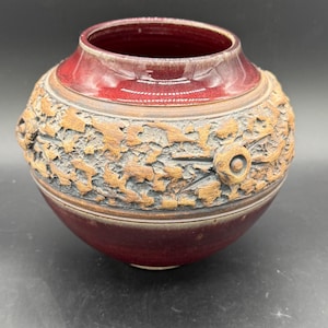 Op de afbeelding: Een handgemaakte, ronde keramische vaas met een dieprode glazuurlaag aan de boven- en onderkant. Het middelste gedeelte heeft een getextureerd, gesneden ontwerp in bruin- en grijstinten, wat een rustieke esthetiek toevoegt. De vaas heeft een brede opening.