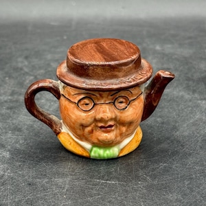 Puede incluir: Una tetera novedosa con forma de cabeza de hombre, con sombrero marrón y gafas. La tetera está pintada en tonos naranja, marrón y verde. Tiene un asa y un pico, y es un objeto de colección peculiar.