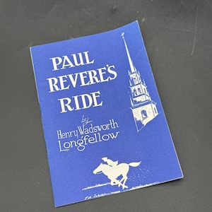 Puede incluir: Un folleto azul titulado "Paul Revere's Ride" de Henry Wadsworth Longfellow. La portada presenta una ilustración blanca de un campanario y un jinete a caballo. El folleto está sobre una superficie oscura.