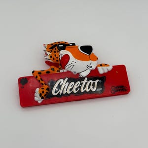 Op de afbeelding: Een rode, rechthoekige Cheetos-magneet met Chester Cheetah, de mascotte, met oranje vacht en zwarte vlekken. De magneet heeft het Cheetos-logo in witte letters.