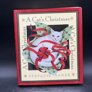 May include: A Cat's Christmas book cover featuring a white cat with yellow eyes, wrapped in a red ribbon and bow. The book has a red border with the title and author's name, Stefanie Samek, displayed.