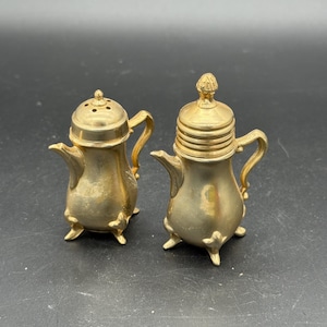 Pode incluir: Dois saleiros e pimenteiros antigos dourados. Cada saleiro e pimenteiro tem a forma de uma pequena chaleira com uma pega e uma tampa. Os saleiros e pimenteiros têm três pés pequenos e são feitos de metal brilhante.