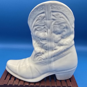 May include: A white ceramic cowboy boot planter featuring embossed decorative stitching and patterns on the shaft and toe. The boot has a rounded heel and a pointed toe, with a glossy finish that highlights the molded details of the design.