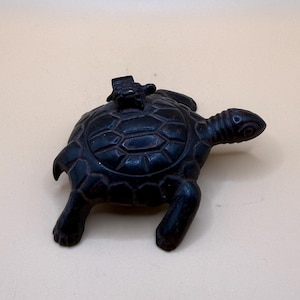 Könnte beinhalten: Eine dunkelbraune, geschnitzte Schildkrötenfigur mit einer kleineren Schildkröte auf ihrem Panzer. Die größere Schildkröte hat ein detailliertes, segmentiertes Panzermuster, vier Beine und einen ausgestreckten Kopf. Die Oberfläche wirkt glatt und matt, mit sichtbaren geschnitzten Linien, die die Panzerplatten und die Gliedmaßen beider Kreaturen definieren.