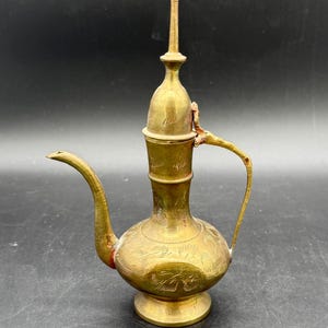 Puede incluir: Una jarra de latón antigua con un pico largo y un asa. La jarra presenta intrincados patrones florales grabados y una tapa decorativa alta. El latón tiene una pátina cálida y envejecida, lo que indica su antigüedad. La jarra probablemente se usa para servir bebidas.