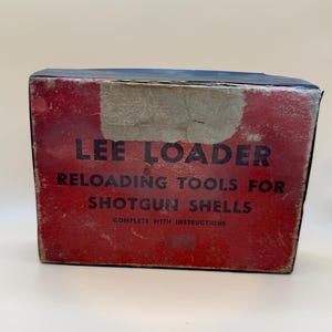 May include: A rectangular box for a Lee Loader reloading tool set for shotgun shells. The box is primarily red with black text that reads LEE LOADER RELOADING TOOLS FOR SHOTGUN SHELLS COMPLETE WITH INSTRUCTIONS. The box shows signs of wear.