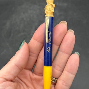 以下が含まれることがあります： Mr.ピーナッツのフィギュアがトップに付いたヴィンテージペン。ペンは青と黄色のバレルで、「Mr.ピーナッツ」の文字が印刷されています。ペンは手で持たれています。